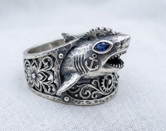 Puede incluir: Anillo de plata con forma de tiburón, con un ojo de piedra azul y detalles intrincados. El anillo presenta un ancla y un diseño floral en la banda. La boca del tiburón está abierta, revelando dientes. Una joya única.