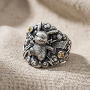 May include: A silver ring featuring a central sculpted character with pointed ears, surrounded by embossed cloud patterns and lightning bolt motifs. The design includes two small yellow circular stones and one clear white stone set into the metal.