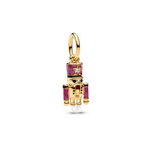 May include: A gold-toned metal charm shaped like a nutcracker soldier. The figure features a glittery pink hat and sleeves, black accents, white boots, and a small clear stone set into a star on the hat. It includes a circular gold jump ring.