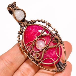May include: A wire-wrapped pendant featuring a large, oval-shaped, faceted ruby-colored gemstone as the centerpiece. The pendant is crafted from copper wire and includes two small, round, translucent white moonstone beads integrated into the design.