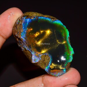 May include: A raw, dark-toned boulder opal featuring a polished, deep black center with vibrant flashes of gold, green, and blue iridescence. The stone has a rough, textured brown outer edge and displays the text OpalByAbdul in the center.