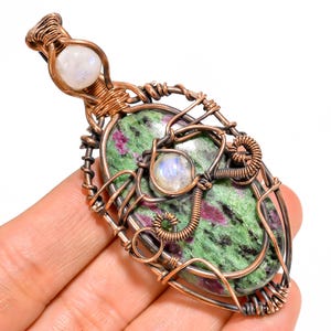May include: An oval-shaped pendant featuring a large green and magenta zoisite stone accented with two round, translucent moonstones. The stones are encased in a decorative, hand-wrapped copper wire frame with intricate coiled details.