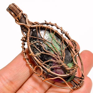 May include: A pendant featuring an oval-shaped dark stone with green and deep red inclusions, encased in a decorative copper wire wrap. The wire is intricately coiled to form a tree-like design over the stone, with a looped bail at the top.