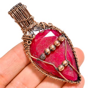 May include: A teardrop-shaped pendant featuring a large, faceted deep red gemstone encased in intricate, hand-woven copper wire. The design includes a decorative bail at the top and a series of small, polished copper beads arranged across the stone.