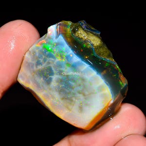 Peut inclure: Une pierre précieuse opale brute et non taillée présentant une base blanc laiteux translucide avec des éclats vibrants de vert, d'orange et de jaune. La pierre présente une surface texturée irrégulière avec des inclusions sombres et le texte OpalByAbdul.