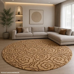 May include: A round, tan-colored area rug featuring a repeating, raised, dark brown floral pattern of stylized, swirling blossoms. The rug is positioned on a light-colored floor. The text RUGSSTUDIOHANDMADE is visible in the bottom left corner.