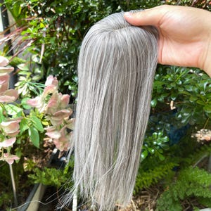 May include: A hair topper featuring straight, shoulder-length strands in a blend of silver, gray, and white tones. The piece has a natural-looking base at the top, designed to provide coverage and volume, with smooth, sleek hair fibers throughout.