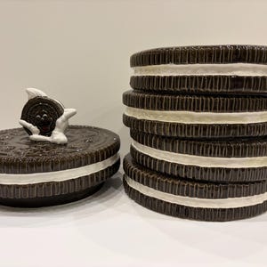 Op de afbeelding: Een keramische koekjespot in de vorm van een Oreo-koekje, met een deksel met een grillig ontwerp. De pot is donkerbruin met een witte crèmevulling en een stapel vergelijkbare koekjes is rechts zichtbaar.