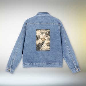 May include: Light blue denim jacket with a graphic print on the back. The print features a surreal illustration of a spinning top, books, and a cliffside under a moonlit sky. The jacket has a classic design with button closures and pockets.