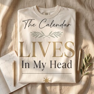 以下が含まれることがあります： クリーム色のクルーネックTシャツ。黒の筆記体と金のセリフ体で「The Calendar LIVES In My Head」という文字がプリントされ、中央下部には緑の葉のイラストと金の星のシンボルがあしらわれています。