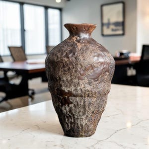 May include: A textured, brown ceramic vase with a wide body and a narrow neck. The vase has a rough, stone-like appearance with darker brown accents. The vase is displayed on a white marble surface.