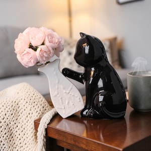 Può includere: Una statuina di gatto nero lucido che tiene un vaso bianco con rose rosa. Il gatto in ceramica ha un design elegante e moderno e il vaso ha una superficie strutturata. La composizione è su un tavolo di legno, creando un'affascinante esposizione decorativa.