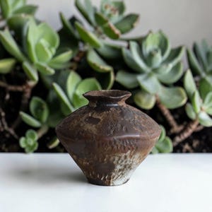 May include: A white ceramic mug with a black handle and the text "But First, Coffee" in a black, handwritten-style font. The mug is on a white surface.