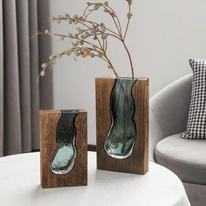 May include: Two decorative vases. Each vase features a dark teal glass insert within a rectangular wooden frame. One vase is taller than the other. A branch with small, clear, bead-like accents is placed in the taller vase. The vases are on a white table.