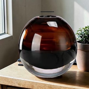 May include: A large, spherical glass vase with a gradient color scheme. The top half is a rich brown, transitioning to a clear base. The vase is sitting on a wooden surface, with a small potted plant visible in the background.