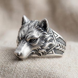 Könnte beinhalten: Ein silberner Ring mit einem detaillierten Wolfskopf-Design. Das Gesicht des Wolfes ist aufwendig modelliert und zeigt Fellstruktur und Gesichtszüge. Das Ringband hat geometrische Muster. Der Ring wird auf einer neutralen, strukturierten Oberfläche präsentiert.
