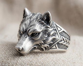 May include: A silver-colored ring featuring a detailed wolf head design. The wolf's face is intricately sculpted, showcasing fur texture and facial features. The ring band has geometric patterns. The ring is displayed on a neutral, textured surface.