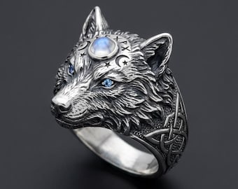 Könnte beinhalten: Ein silberfarbener Ring mit einem detaillierten Wolfskopf-Design. Der Wolf hat blaue Edelsteinaugen und einen Mondstein auf der Stirn. Der Ring hat ein kompliziertes keltisches Knotenmuster.