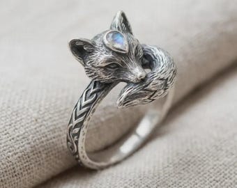 May include: A silver fox ring with a detailed fox head and tail design. The fox's head features a teardrop-shaped, iridescent gemstone. The ring band has an etched pattern. A unique piece of jewelry.