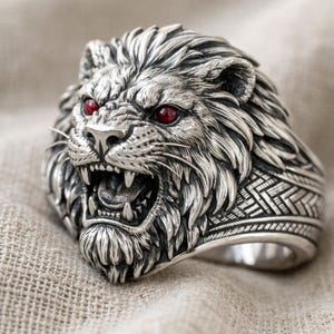Può includere: Anello a forma di leone color argento con occhi di pietra rossa. L'anello presenta una testa di leone dettagliata con la bocca aperta, che mostra denti affilati. La fascia dell'anello ha un intricato motivo geometrico.