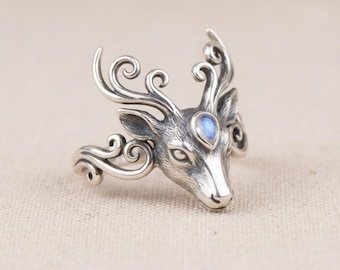 May include: A silver-toned ring featuring a deer head design. The ring has intricate detailing on the antlers and face, with a teardrop-shaped, blue-hued stone set between the antlers. The ring's band has a swirling pattern.