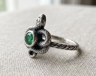 May include: A silver ring featuring a coiled snake design. The snake wraps around a round, vibrant green gemstone. The ring's band has a textured pattern. The ring is set against a neutral background.