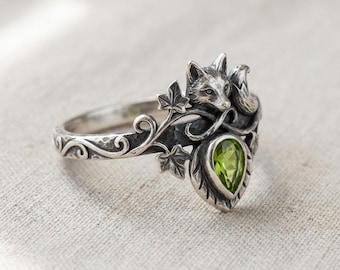 May include: A silver ring featuring a fox design with a green pear-shaped gemstone. The ring has intricate details, including leaves and swirling patterns. The fox is sculpted on the top of the ring.
