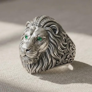 May include: Silver lion ring with green gemstone eyes and a detailed mane. The ring features a decorative design on the band and small clear stones on the lion's head. A statement piece of jewelry.