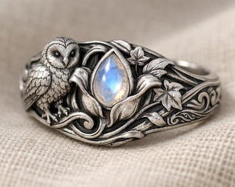 May include: A silver-toned ring featuring an owl and a teardrop-shaped, iridescent gemstone. The ring has intricate floral and leaf detailing around the gemstone and along the band. The owl is perched on the side of the ring, adding a whimsical touch.