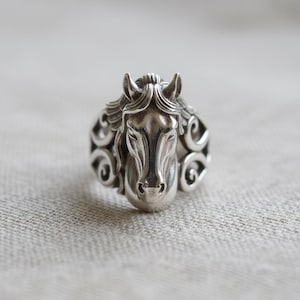 May include: A silver ring featuring a detailed horse head design. The horse's mane and face are intricately sculpted, with decorative swirls on the ring's band. The ring is made of a shiny, metallic material.