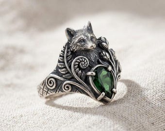 May include: A detailed silver ring featuring a fox head and foliage design. The ring has a pear-shaped, emerald-green gemstone at its center. The ring's design includes intricate details and a textured finish, showcasing a nature-inspired theme.