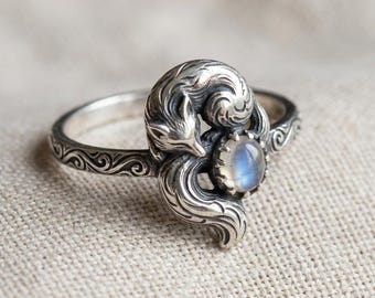 May include: A silver ring featuring a detailed fox design curled around a blue-hued gemstone. The ring band has an ornate, swirling pattern. The fox's fur and features are intricately carved, creating a whimsical and elegant piece of jewelry.