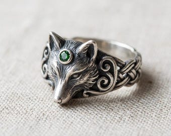 May include: A silver ring featuring a detailed wolf head with a green gemstone eye. The ring has a braided band design and intricate scrollwork on the sides, creating a unique and detailed piece of jewelry.