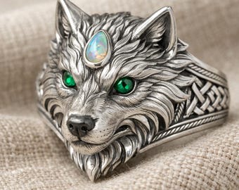 May include: A detailed silver ring featuring a wolf's head with green eyes and an opal stone on its forehead. The ring has intricate detailing on the wolf's fur and a Celtic knot design on the band.