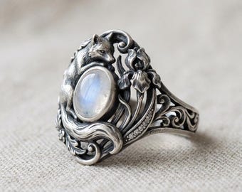 May include: A silver ring featuring a fox and floral design. The ring has an oval, iridescent gemstone at its center. The fox is sculpted on the side, with floral and scrollwork details. Small clear stones are set along the band.