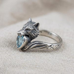 Peut inclure: Bague en argent représentant une tête de loup tenant une pierre précieuse bleue clair en forme de cœur. La bague présente des détails fins sur la fourrure du loup et un motif texturé sur l'anneau. Un bijou unique.