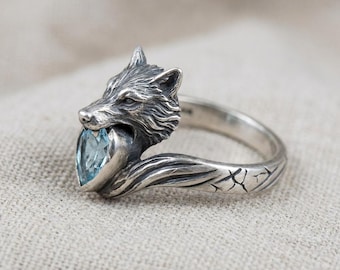 May include: A silver ring featuring a wolf head biting a heart-shaped, light blue gemstone. The ring has intricate details on the wolf's fur and the band. The band has a textured design. A unique piece of jewelry.