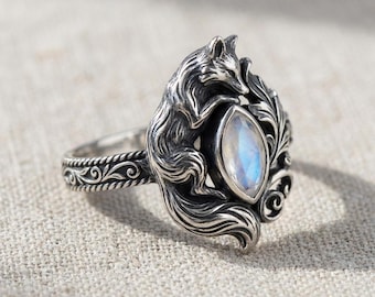 May include: A silver ring featuring a detailed fox design. The fox is sculpted around a pear-shaped, iridescent gemstone. The ring band has an ornate, rope-like pattern. The ring is set against a neutral background.