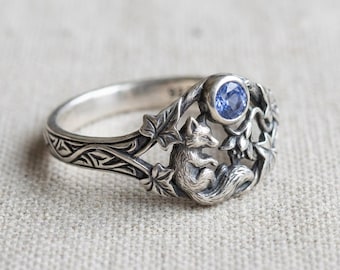 May include: A silver ring featuring a fox nestled among leaves and a blue gemstone. The ring has intricate detailing, with a fox design and leaf accents. The gemstone is round and a vibrant blue color.