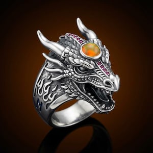 May include: A silver dragon ring with an orange gemstone and small red stones. The dragon has detailed scales, horns, and an open mouth. The ring features a flame design on the side.
