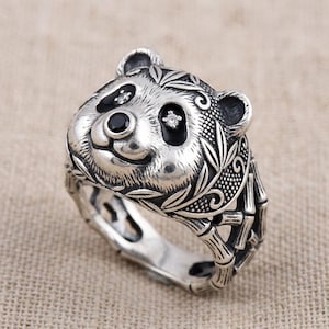 May include: A silver-colored ring featuring a panda bear design. The panda's face has black eyes and a black nose, with a small gem in each eye. The ring band is decorated with bamboo stalks and leaves.