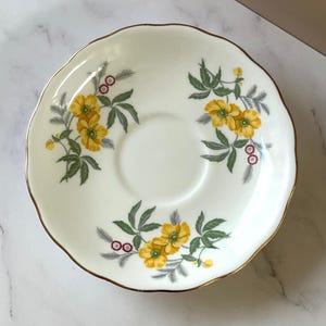 May include: A white saucer with a scalloped edge and a gold rim. The saucer is decorated with yellow flowers, green leaves, and small red berries. A classic design for a tea set.