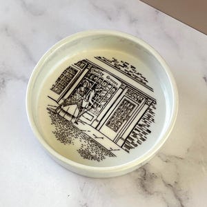 May include: A round, off-white ceramic dish with a black and white illustration of a shop with the words "Quality Grocers." The dish has a slightly raised edge and a smooth surface.