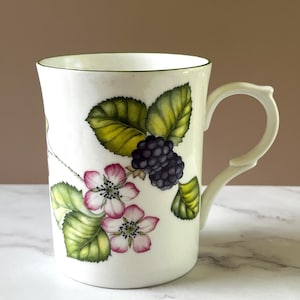 May include: White ceramic mug with a green rim and handle. The mug features a botanical design of blackberries, pink flowers, and green leaves. The handle has a distinctive, curved shape.