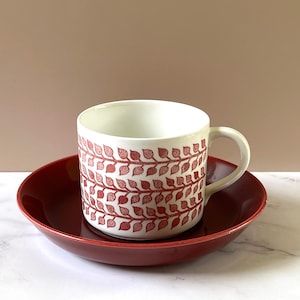 May include: A white ceramic mug with a red leaf pattern sits on a matching red saucer. The mug has a curved handle and the leaf design covers the exterior. The saucer is round and slightly concave, creating a cohesive set.
