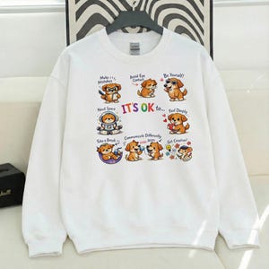 May include: White sweatshirt with a colorful graphic featuring cartoon dogs. The design includes the text "IT'S OK to..." with various phrases like "Make Mistakes" and "Be Yourself!" The sweatshirt has a crew neck and long sleeves.