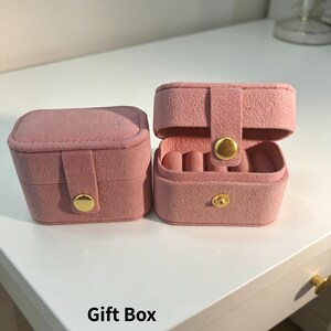 May include: Two pink velvet jewelry boxes, one closed and one open, displaying ring holders. Both boxes have gold-colored clasps. The text "Gift Box" is at the bottom of the image.