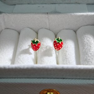 May include: A pair of strawberry-shaped stud earrings. Each earring features a red enamel strawberry with green leaves and gold-colored metal accents. The earrings are displayed in a white jewelry box.