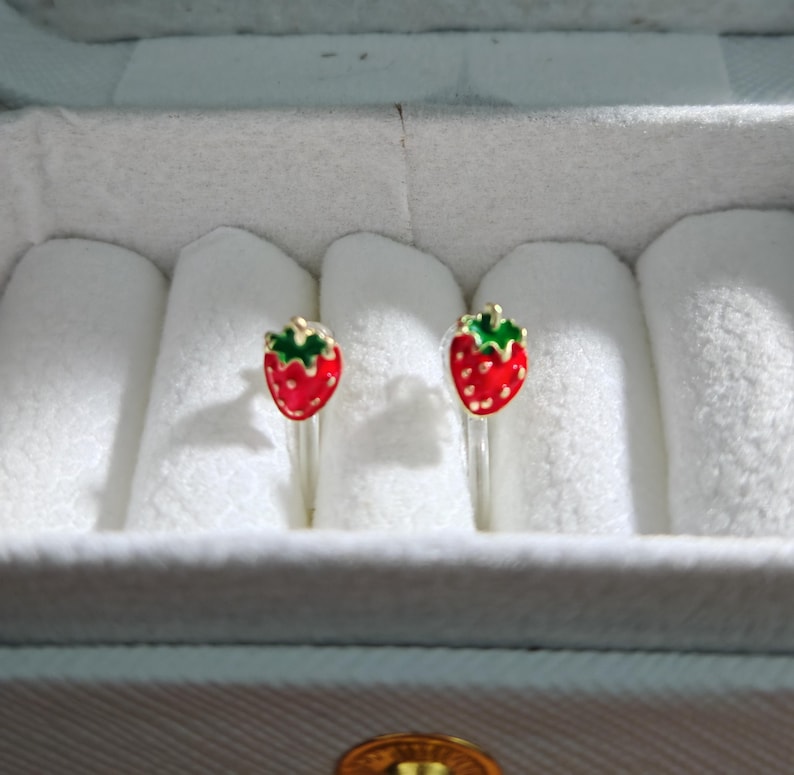 May include: A pair of strawberry-shaped stud earrings. Each earring features a red enamel strawberry with green leaves and gold-colored metal accents. The earrings are displayed in a white jewelry box.