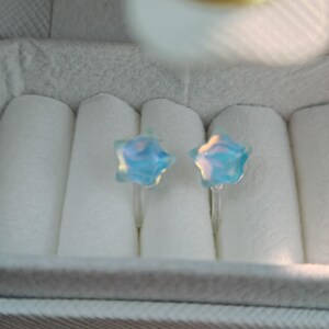 May include: A pair of star-shaped earrings with a light blue, iridescent finish. The earrings are displayed in a white jewelry box with a gray and white striped exterior. The earrings are small and delicate.